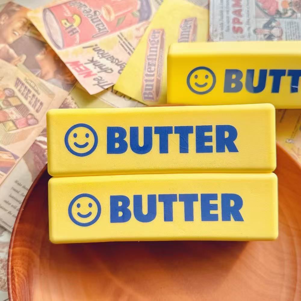 Two blue-on-yellow smiley-face BUTTER squishy sticks stacked on a wooden plate over vintage newspaper