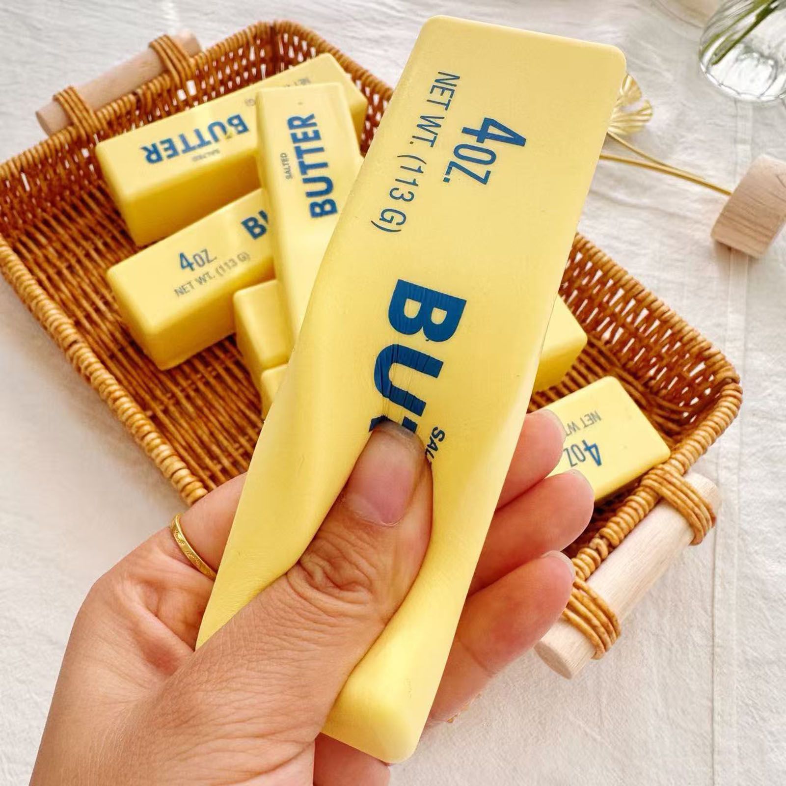 A hand holding a yellow butter stick squishy over a woven basket full of them